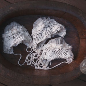 May include: Three white crocheted baby bonnets with delicate lace trim, arranged in a wooden bowl.