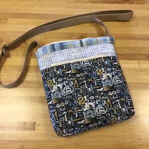 May include: A small black crossbody bag with a brown strap. The bag is made of fabric with a pattern of blue, yellow, and white letters and symbols. The bag has a zippered pocket on the front.