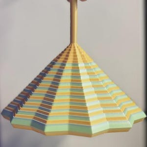 May include: A colorful, geometric-patterned toy top with a yellow handle. The top features alternating stripes of yellow, green, and blue, with brown accents. The base has a multi-faceted design. The toy is held by a hand.