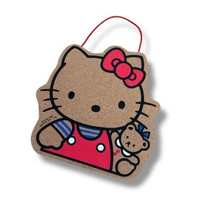 May include: A cork wall hanging shaped like Hello Kitty, featuring a red bow, red overalls, and a small teddy bear. The character has black whiskers, eyes, and a nose. A red string is attached to the top. Text reads 1976, 1995 SANRIO CO., LTD.