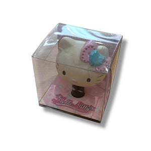 May include: A Hello Kitty bottle stopper in a clear plastic box. The stopper features the iconic character's face with a pink bow and blue heart. The packaging has a pink background with the words "Hello Kitty".