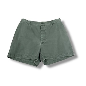 Könnte beinhalten: Eine grüne Shorts in Salbeigrün. Die Shorts haben einen Knopf- und Reißverschluss, Gürtelschlaufen und zwei Vordertaschen. Die Shorts sind aus einem robusten Stoff gefertigt und haben einen klassischen, lässigen Stil.