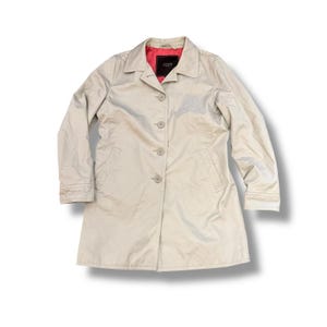 May include: A beige button-down coat with a collar and long sleeves. The coat has a red interior lining and a small black label. The coat is a neutral color and has a classic design.