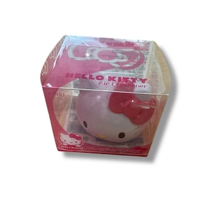 May include: A Hello Kitty air freshener in a clear plastic box. The air freshener is shaped like Hello Kitty's head, with a pink body and a red bow. The box has pink and white accents and the words "HELLO KITTY Air Freshener".