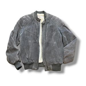 May include: A grey suede bomber jacket featuring a cream-colored faux shearling lining, a front zipper closure, ribbed cuffs and hem, quilted shoulder panels, and two side pockets. The jacket has a stand-up collar with a snap button closure.