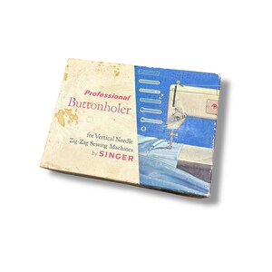 Puede incluir: Caja vintage Singer Professional Buttonholer. La caja es beige y azul, con las palabras "Professional Buttonholer" en rosa y el logotipo de Singer. La caja muestra una imagen de una máquina de coser.