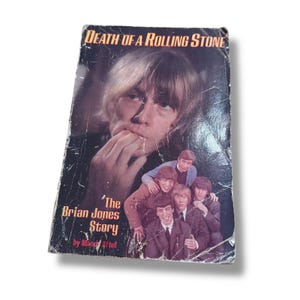 以下が含まれることがあります： 「Death of a Rolling Stone」というタイトルのヴィンテージ本の表紙。男性の肖像画と男性のグループが描かれています。表紙には「The Brian Jones Story」というテキストと著者の名前も含まれています。本は使い古された外観をしています。