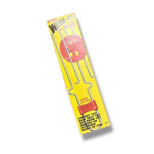May include: Vintage "Whee-Lo" toy in its original packaging. The yellow box features the toy's name and a red wheel design. The toy itself is visible through the packaging, with a red wheel and metal frame. Text on the box reads "Contest Toy" and "Hours of Fun."