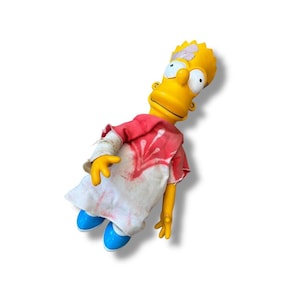 Puede incluir: Muñeco amarillo de Bart Simpson con camisa roja y blanca y zapatos azules. El muñeco tiene una venda en el brazo y un parche en la cabeza. Un artículo de colección.