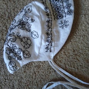 May include: White linen bonnet with black embroidered floral pattern and white ties.