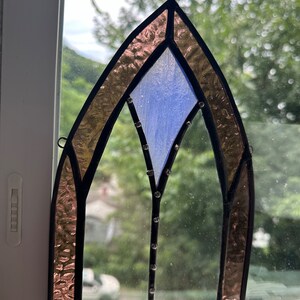 May include: A stained glass window with a pointed arch design. The window is made of clear glass with a blue diamond shape in the center. The edges of the window are framed in black and the glass is surrounded by a border of pink glass.