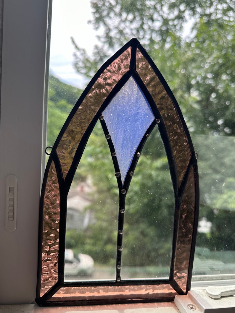 May include: A stained glass window with a pointed arch design. The window is made of clear glass with a blue diamond shape in the center. The edges of the window are framed in black and the glass is surrounded by a border of pink glass.