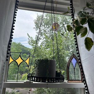 May include: A hanging planter with a chain and a black metal frame. A small potted plant hangs from the planter. The planter is in front of a window with stained glass panels and a view of trees.