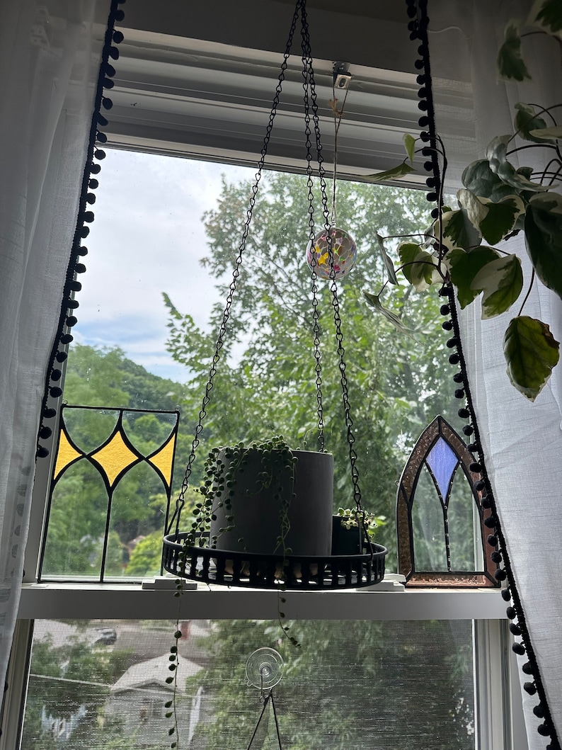 May include: A hanging planter with a chain and a black metal frame. A small potted plant hangs from the planter. The planter is in front of a window with stained glass panels and a view of trees.
