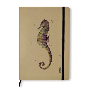 Puede incluir: Una libreta marrón con una ilustración de acuarela de un caballito de mar en la portada. La libreta tiene un cierre elástico negro y el texto "JULIE RO" está impreso en la esquina inferior derecha.