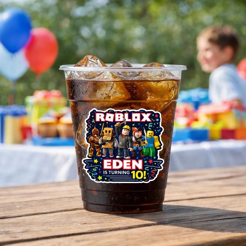 May include: A clear plastic cup filled with a dark beverage and ice cubes. A colorful sticker on the cup features the Roblox logo and the text "Eden is turning 10!" Balloons and party decorations are visible in the background.