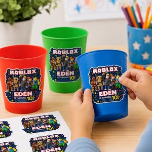 May include: Three plastic cups in red, green, and blue, each featuring a Roblox sticker with the text "ROBLOX EDEN IS TURNING 10!". A sheet of matching stickers is visible on the table, and a blue pencil holder with white stars sits in the background.