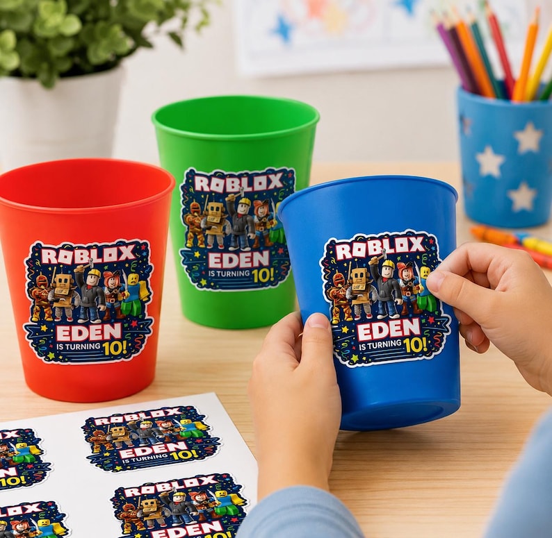 May include: Three plastic cups in red, green, and blue, each featuring a Roblox sticker with the text "ROBLOX EDEN IS TURNING 10!". A sheet of matching stickers is visible on the table, and a blue pencil holder with white stars sits in the background.