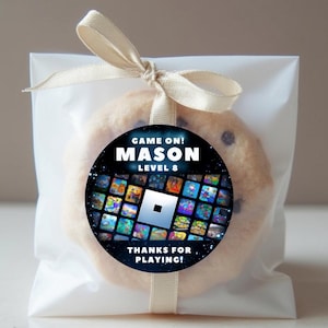 May include: A chocolate chip cookie in a clear cellophane bag, tied with a beige ribbon. A round sticker on the cookie reads "GAME ON! MASON LEVEL 8" with a Roblox theme and the words "THANKS FOR PLAYING!"