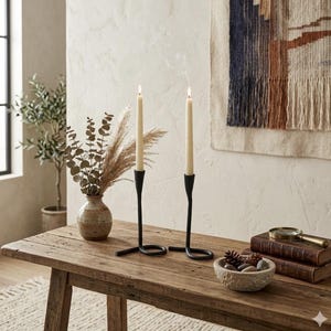 Peut inclure: Deux bougies fines et allumées, de couleur crème, dans des bougeoirs en métal noir avec une base en boucle. Les bougeoirs sont sur une table en bois rustique, à côté d'un petit vase avec des plantes séchées et un bol décoratif.