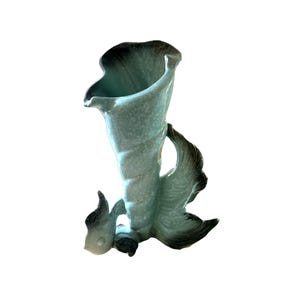 May include: A light green ceramic vase in the shape of a fish. The vase has a flared opening and a textured surface. The fish tail and fins are a darker shade of green.