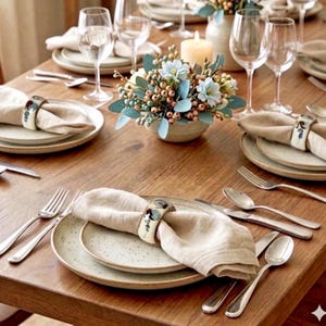 Peut inclure: Une table à manger en bois dressée avec des assiettes en céramique mouchetée, des serviettes en lin beige retenues par des ronds de serviette en céramique à motifs, des couverts en argent, des verres à vin et un centre de table floral.