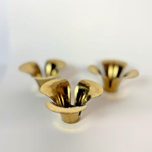 Könnte beinhalten: Drei goldfarbene, blütenförmige Kerzenhalter. Jeder Halter hat ein ausgestelltes, blütenblattartiges Design und einen kleinen, kreisförmigen Sockel. Die Kerzenhalter sind aus glänzendem, reflektierendem Metall gefertigt.