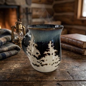 Puede incluir: Una taza de cerámica con un diseño único en tonos azul oscuro y blanco. La taza tiene un asa y un patrón de silueta de bosque. Sale vapor de la taza, lo que sugiere una bebida caliente. La taza está sobre una superficie de madera.