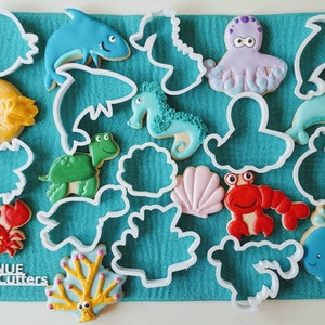 May include: A set of 12 white plastic cookie cutters shaped like various sea creatures, including a crab, a turtle, a starfish, a seahorse, a dolphin, an octopus, a whale, and a seashell. The cutters are arranged on a turquoise blue fabric background. The text "ECINUE Cookie Cutters" is visible in the lower left corner of the image.