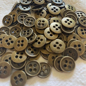 May include: A collection of antique brass-coloured buttons, each with four holes. The buttons are circular with a slightly raised edge. They are scattered on a white surface.
