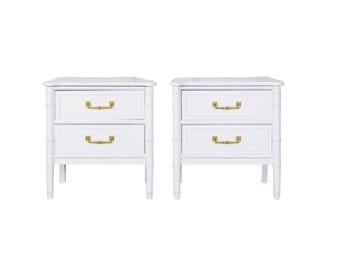 May include: Two white nightstands with two drawers each, featuring gold-colored handles. The nightstands have a bamboo-style design and are set against a white background. Each nightstand has a rectangular top.