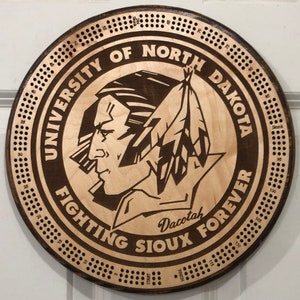 May include: A wooden circular game board with a brown background and a white center. The center features a logo for the University of North Dakota Fighting Sioux. The logo is a Native American profile with a feather headdress. The board is numbered from 1 to 120 around the edge.