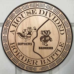 May include: A round wooden cribbage board with the text "A House Divided Border Battle". The board features etched images of a gopher and a badger, with the words "Gophers" and "Badgers" below them. The board has a dark brown border and is ready for play.