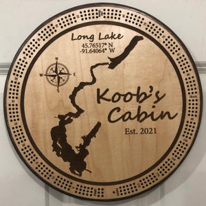 May include: A wooden circular game board with a map of Long Lake, a compass, and the text "Koob's Cabin Est. 2021". The board has a numbered track around the edge.