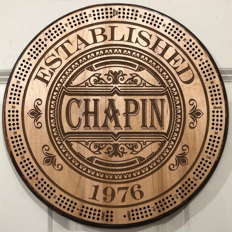 May include: A wooden circular game board with a brown stain. The board has a decorative design with the text "ESTABLISHED CHAPIN 1976". The board has a numbered track around the outside edge.