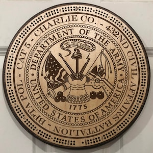 May include: A wooden round plaque with a brown background and a circular design. The design features a seal with the words "Department of the Army" and "United States of America" surrounding a shield with a cannon, a musket, and a flag. The plaque also has the words "Charlie Co. - 432nd Civil Affairs Battalion BPTA POL - CAT 5" and "1775" around the seal.