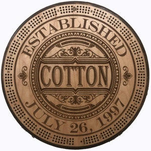 May include: A wooden round sign with a brown background and a decorative border. The sign reads "Established July 26, 1997" and "Cotton" in a decorative font.