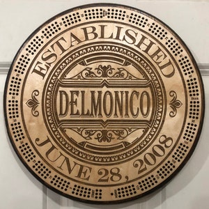 May include: A wooden round plaque with a brown background and a decorative design. The plaque has the text "ESTABLISHED JUNE 28, 2008" and "DELMONICO" in a decorative font.