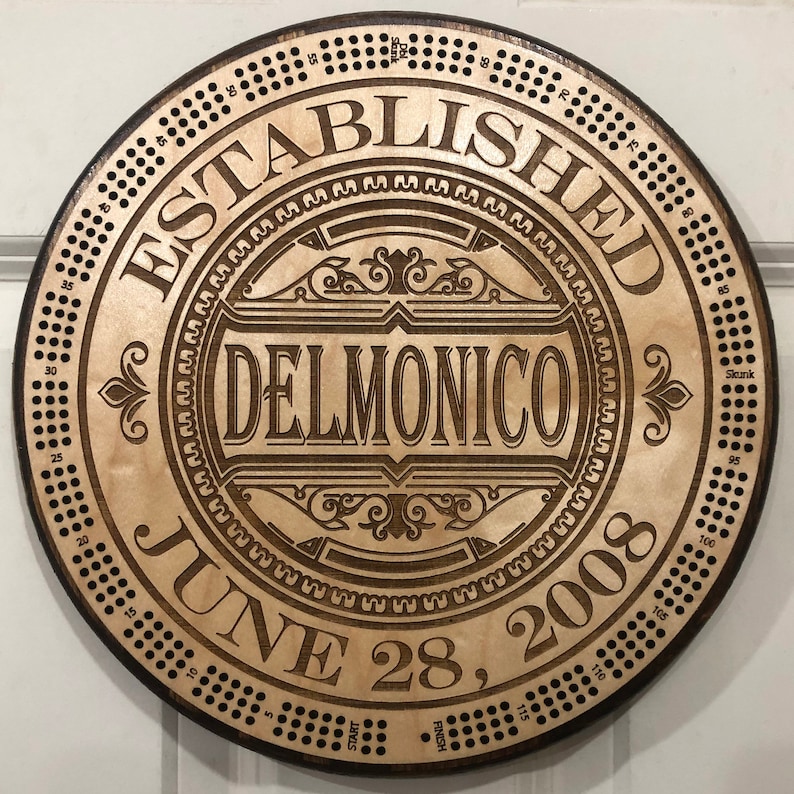 May include: A wooden round plaque with a brown background and a decorative design. The plaque has the text "ESTABLISHED JUNE 28, 2008" and "DELMONICO" in a decorative font.