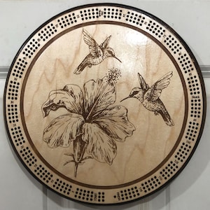 May include: A wooden cribbage board with a brown border and a light wood center. The center features a laser-engraved design of two hummingbirds and a hibiscus flower. The board has a numbered scoring track around the edge.