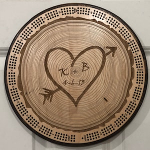 May include: A wooden cribbage board with a heart and arrow design carved into the center. The heart contains the initials "K B" and the date "4-6-13". The board is circular, with a dark brown border and holes for pegs.