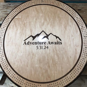 May include: A wooden circular game board with a brown border and black dots around the edge. The center of the board features a mountain range graphic with the text "Adventure Awaits" and the date "5.31.24".