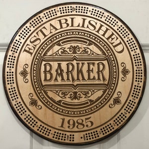 May include: A circular wooden sign with a brown woodgrain finish. The sign features a decorative design with the text "ESTABLISHED BARKER 1985". The sign also has a circular pattern of holes around the edge.