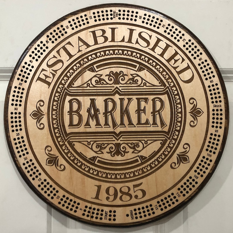 May include: A circular wooden sign with a brown woodgrain finish. The sign features a decorative design with the text "ESTABLISHED BARKER 1985". The sign also has a circular pattern of holes around the edge.