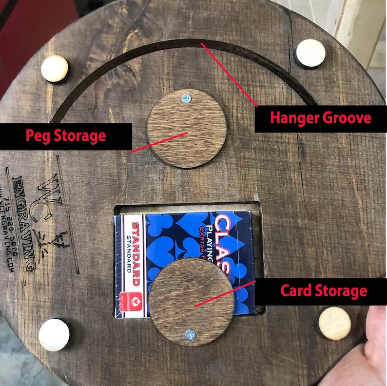 May include: A wooden game board with a groove for hanging, peg storage, and a card storage compartment. The board is made of dark wood and has a natural finish. The text "WC Engraving" is printed on the board.