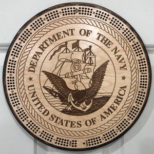 May include: A wooden cribbage board with a circular design featuring the Department of the Navy seal. The seal is a brown eagle with a ship and anchor in the background. The board has a rope design around the edge and numbers from 1 to 120 around the outer edge.
