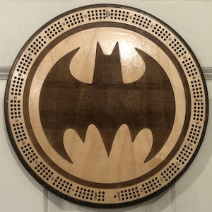 May include: A wooden cribbage board with a Batman logo in the center. The board has a dark brown stain and a light wood finish. The numbers 5 to 120 are printed around the edge of the board.