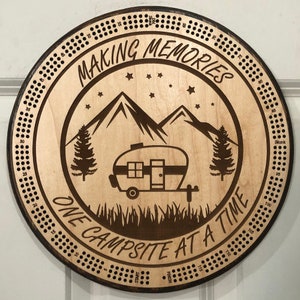 May include: A round wooden cribbage board with a camping theme. The center features a camper, mountains, and trees, with the text "MAKING MEMORIES ONE CAMPSITE AT A TIME." The board has holes for pegs and a dark stained border.
