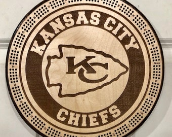 May include: A wooden round game board with a brown background and a white circle in the center. The white circle has the text "KANSAS CITY CHIEFS" in a black font and a red and white arrowhead logo with the letters "KC" in the center. The board is surrounded by a ring of numbers from 1 to 100.