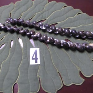 May include: A dark gray beaded necklace with white shell accents. The necklace is displayed on a green leaf-shaped placemat. A white card with the number 4 is also visible.